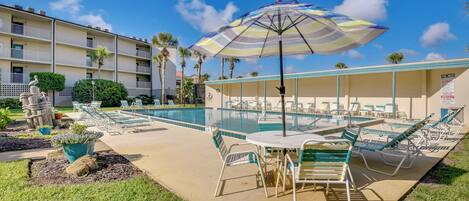 A pool area with lounge chairs and a table, surrounded by a building with balconies and palm trees.