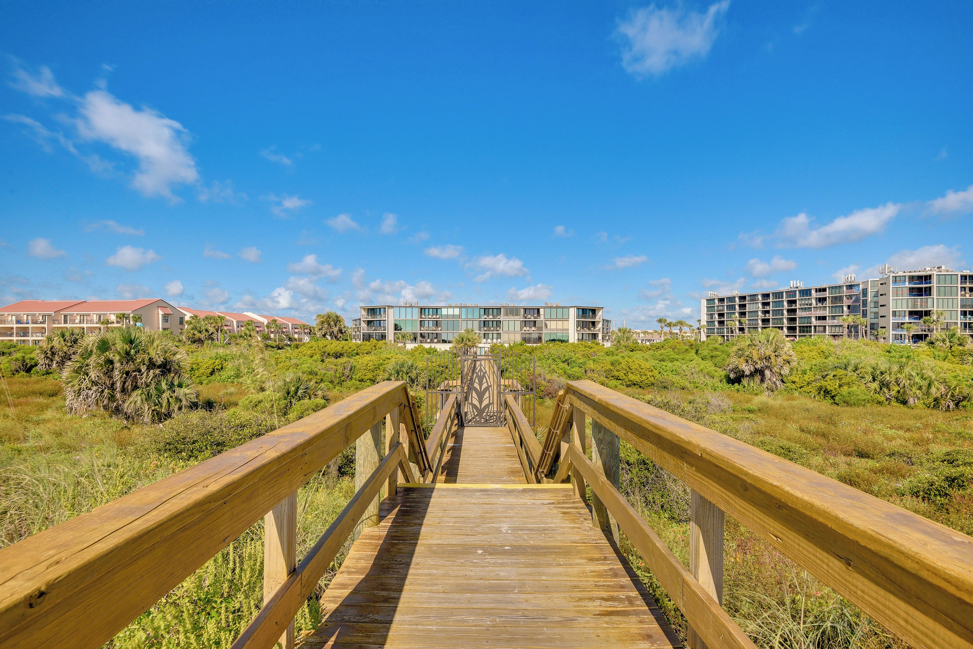 A wooden boardwalk leading to a modern building with multiple floors and balconies, surrounded by greenery.