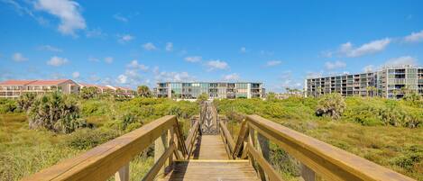 A wooden boardwalk leading to a modern building with multiple floors and balconies, surrounded by greenery.