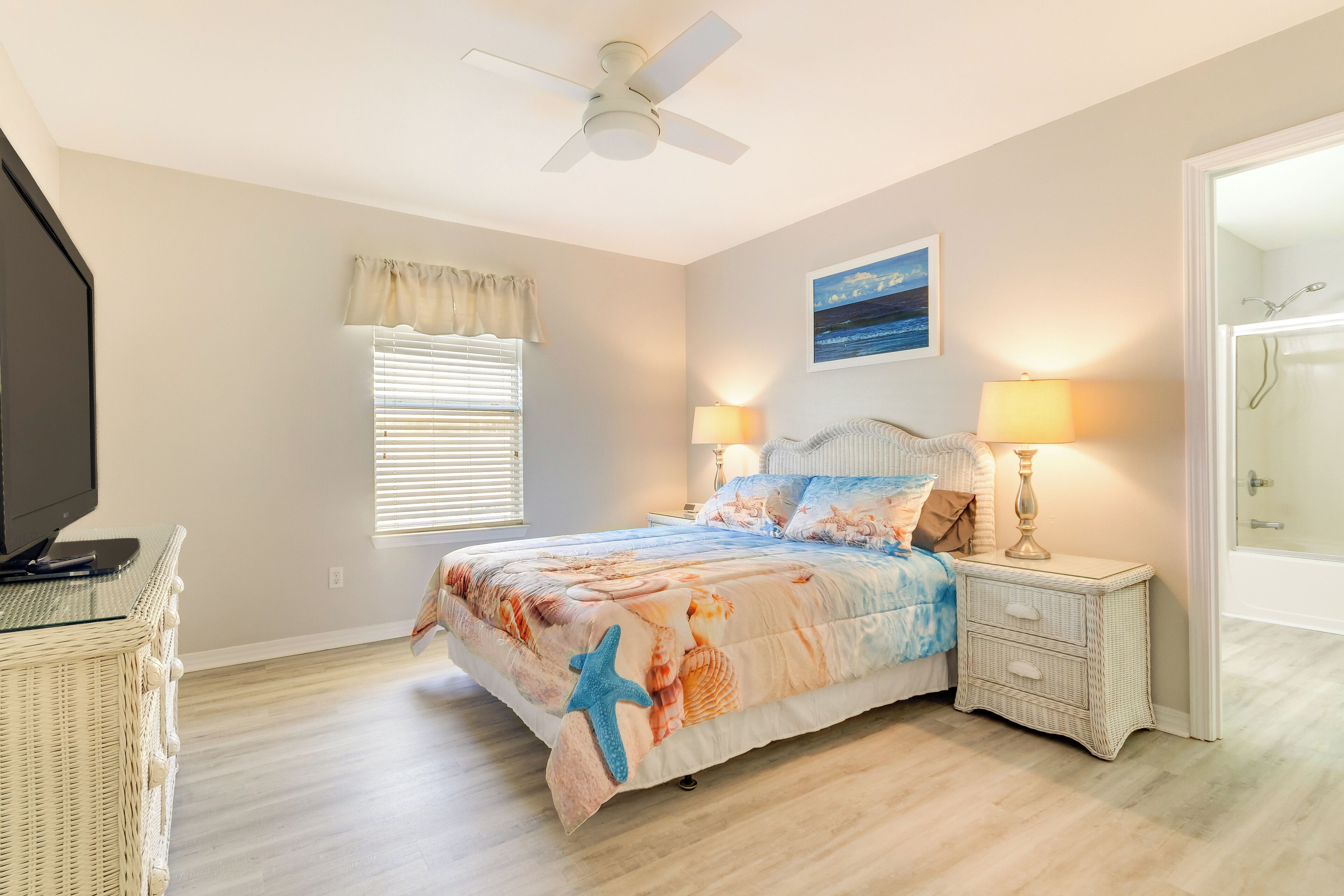 A bedroom with a bed, a ceiling fan, a window with blinds, a nightstand with a lamp, and a television on a stand.