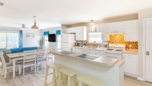 A modern kitchen with white cabinetry, a central island with stools, a dining area with a blue sofa, and a TV mounted on the wall. - Fenced & Private Backyard: Navarre Retreat (Navarre)