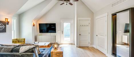 A spacious living room with a high ceiling, a ceiling fan, a large flat-screen TV, a sofa, a coffee table, and a door leading to another room.