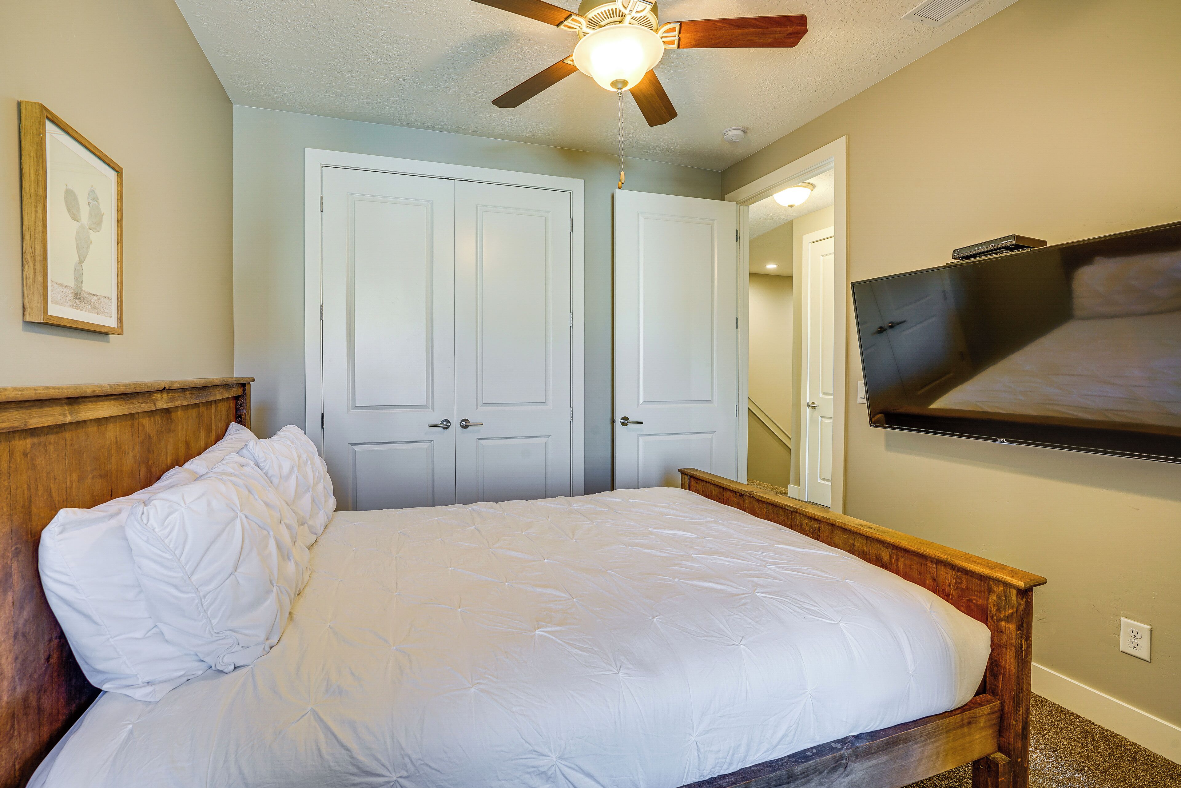 A bedroom with a wooden bed frame, white bedding, a ceiling fan, and a TV mounted on the wall.
