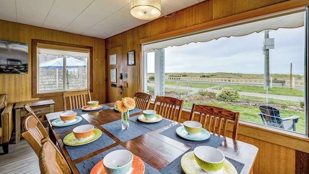 A dining area with a wooden table set for six, chairs, and a large window offering a view of the landscape. - Beachfront Cottage on Seaside Promenade w/ Hot Tub (Seaside)