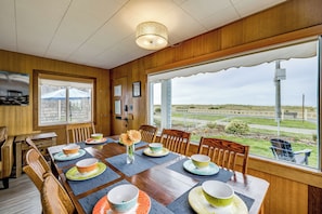 A dining area with a wooden table set for six, chairs, and a large window offering a view of the landscape. - Beachfront Cottage on Seaside Promenade w/ Hot Tub (Seaside)