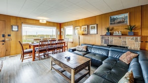 A spacious living room with a large leather sofa, wooden dining table, and chairs. There is a fireplace with a stone mantle and potted plants. - Beachfront Cottage on Seaside Promenade w/ Hot Tub (Seaside)