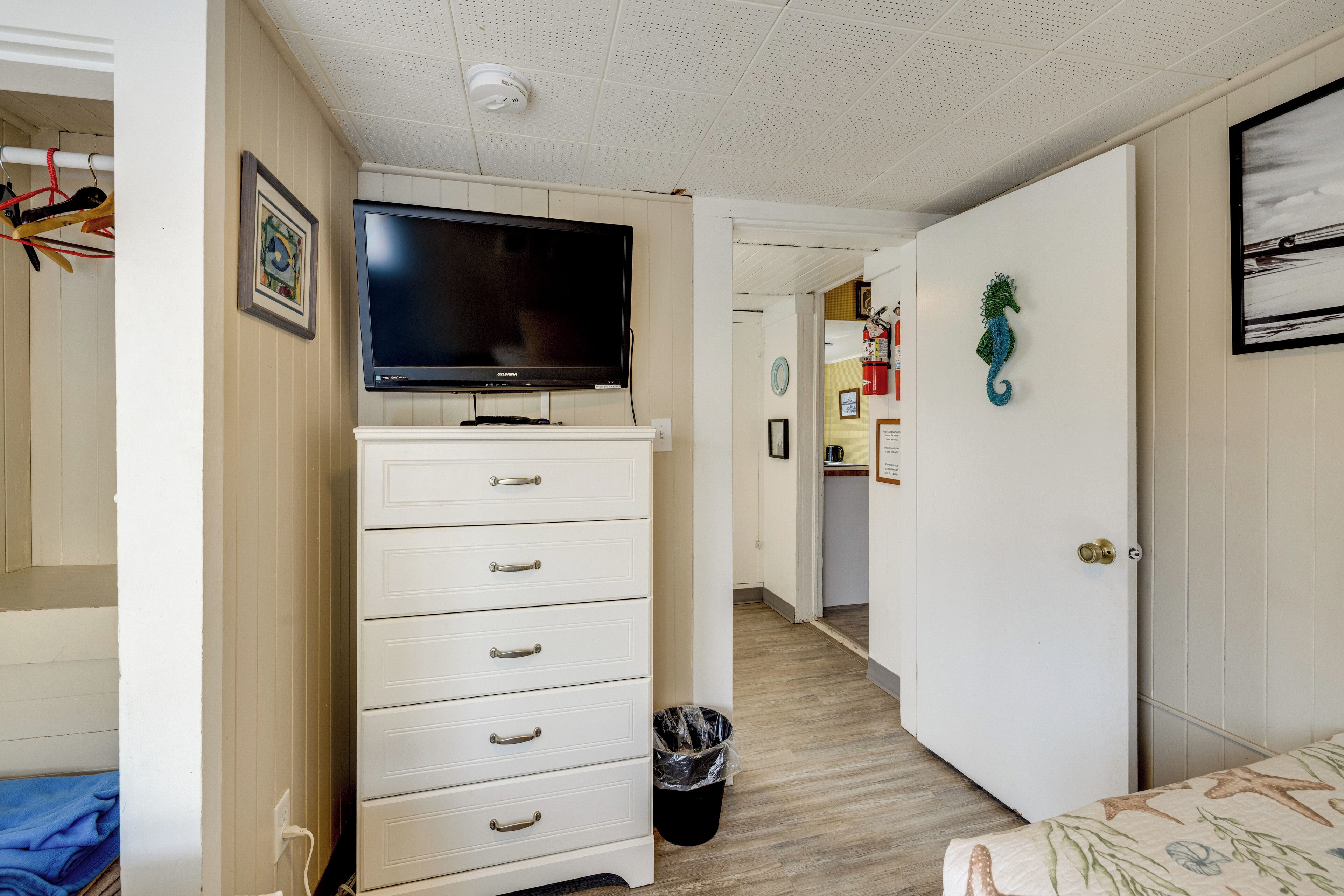 Une chambre à coucher avec un téléviseur posé sur une commode, une porte décorée d’un motif représentant un hippocampe, et un couloir équipé d’un extincteur.