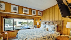 A bedroom with a large bed, a window with a view of parked cars, wooden paneling, and framed pictures on the wall. - Beachfront Cottage on Seaside Promenade w/ Hot Tub (Seaside)