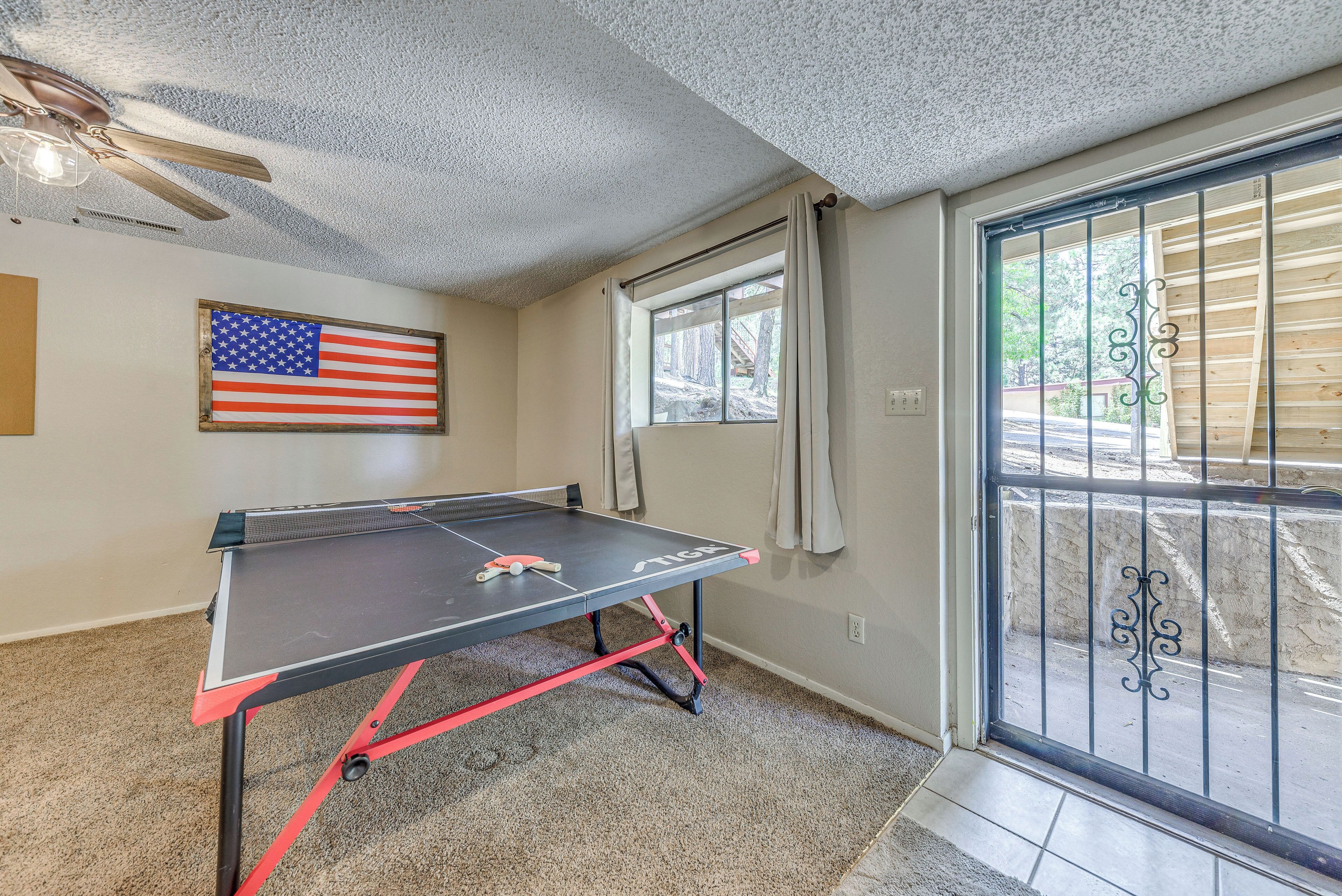 A ping pong table in a room with a ceiling fan, a window with curtains, and a view of a wooden deck outside.