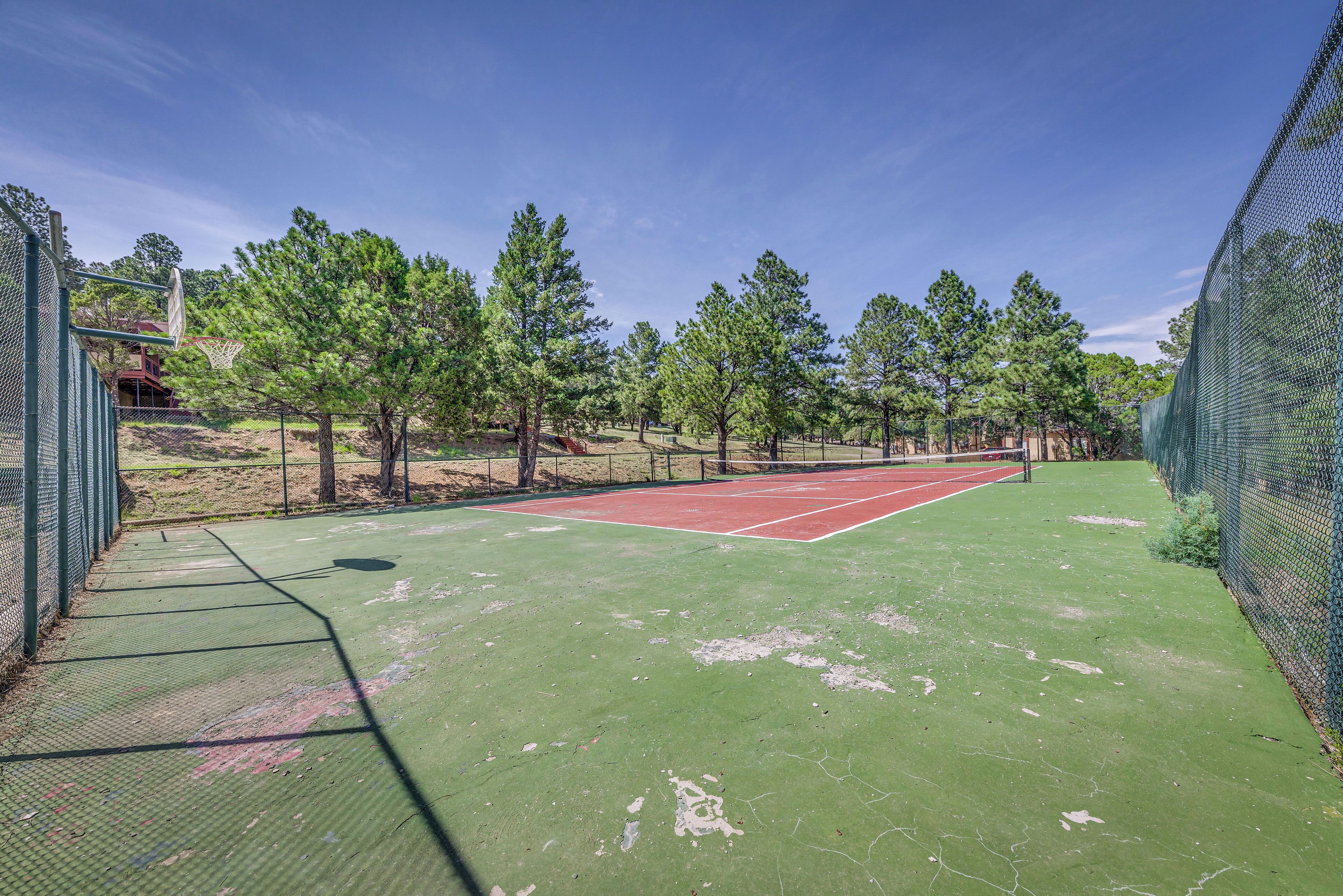 A tennis court with a green surface and a red area, surrounded by trees and a fence.