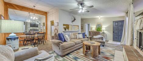 A spacious living room with a beige sofa, a coffee table, and a patterned rug. There is a dining area with a wooden table and chairs, and a kitchen in the background.