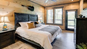 A bedroom with a wooden ceiling, a bed with white bedding, two bedside lamps, a guitar on the wall, and a window with blinds. - Cobbly Nob Resort Cabin: Hot Tub, Pool & Views! (Gatlinburg)