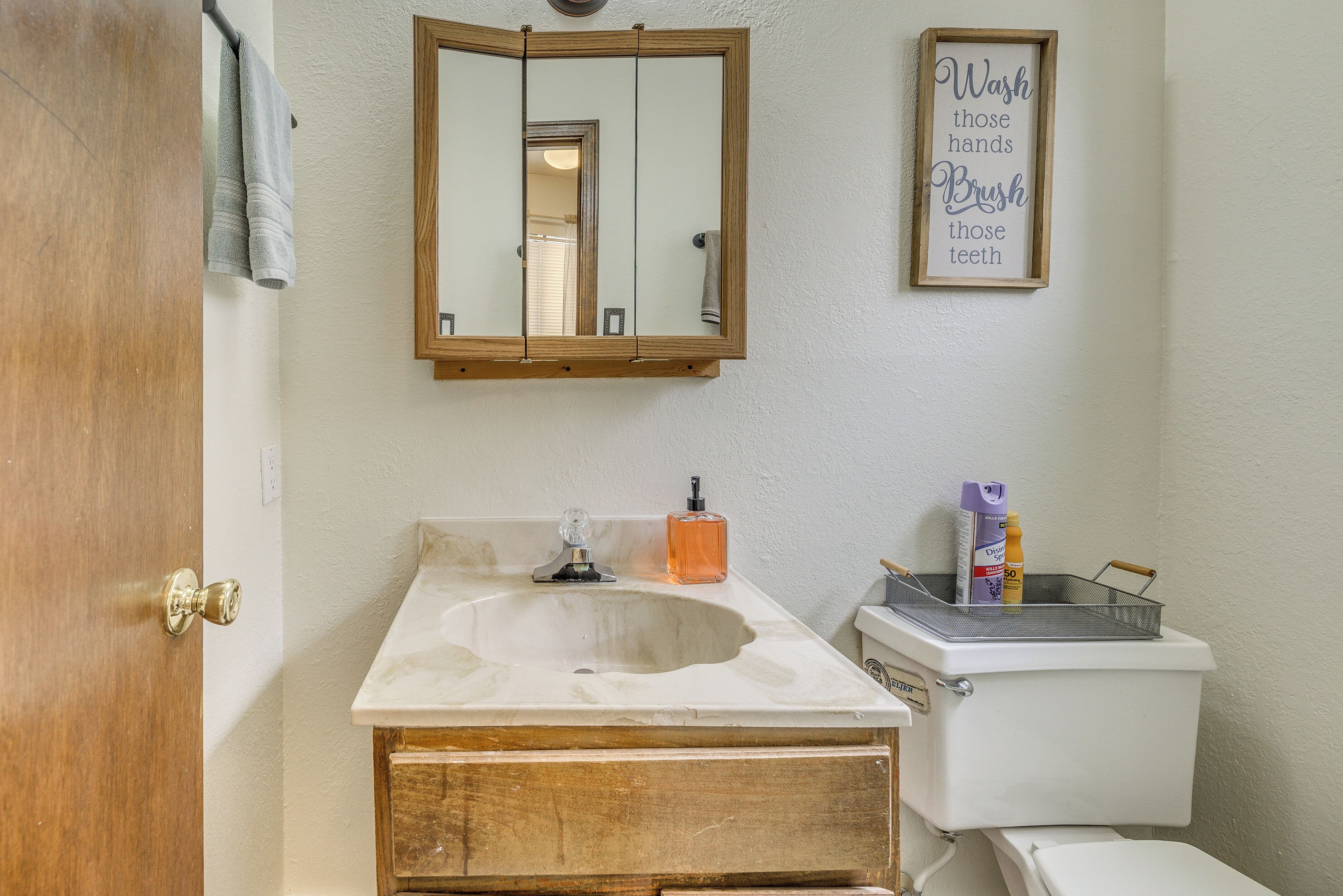 A bathroom with a sink, mirror, and toilet. There is a towel hanging on the door, a framed sign on the wall, and toiletries on the sink.