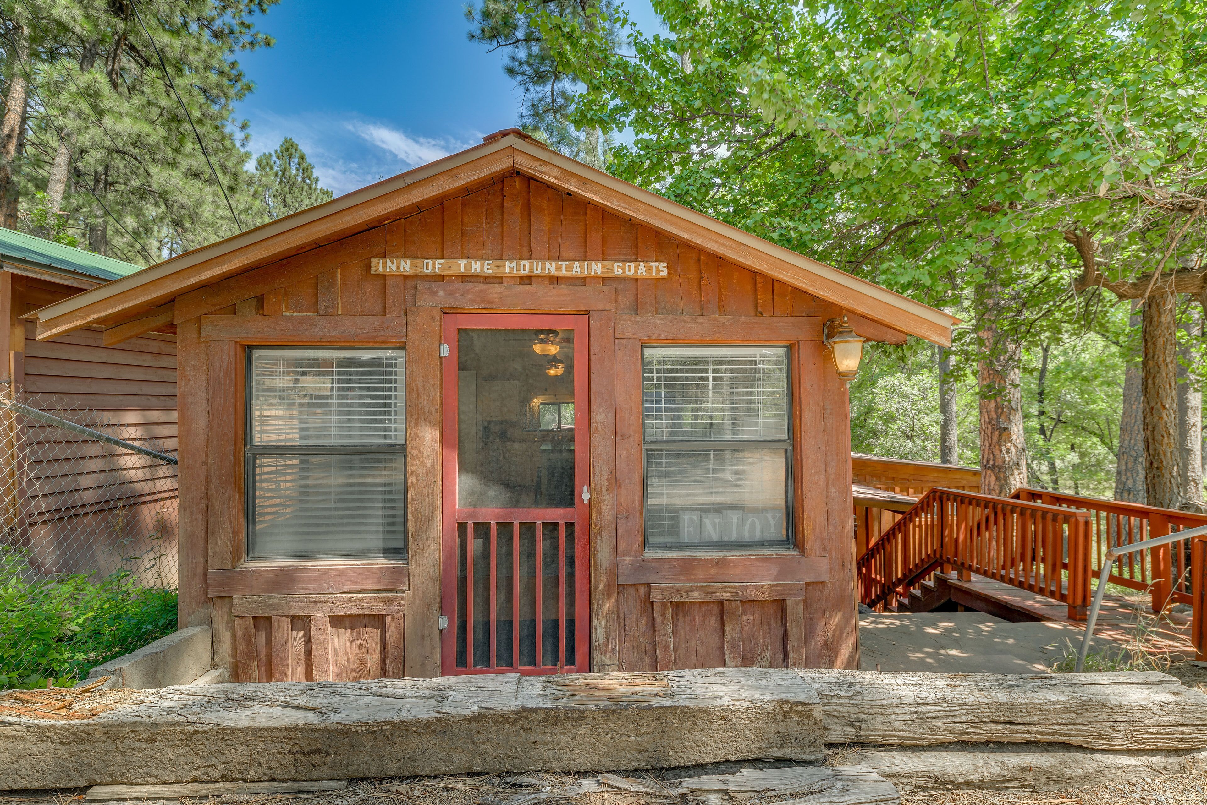 Una cabaña de madera con puerta y ventanas rojas, con el letrero "INN OF THE MOUNTAIN GOATS", rodeada de árboles.
