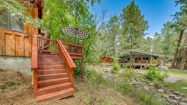 A wooden cabin with a staircase leading to a deck, surrounded by trees and a small stream. - Inn of the Mountain Goats Ruidoso Cabin w/ Deck! (Ruidoso)