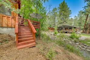 A wooden cabin with a staircase leading to a deck, surrounded by trees and a small stream. - Inn of the Mountain Goats Ruidoso Cabin w/ Deck! (Ruidoso)
