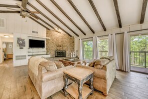 A spacious living room with a stone fireplace, wooden beams, a large TV, and a balcony with curtains. - Pet-friendly Ruidoso Escape w/ Spacious Deck! (Ruidoso)