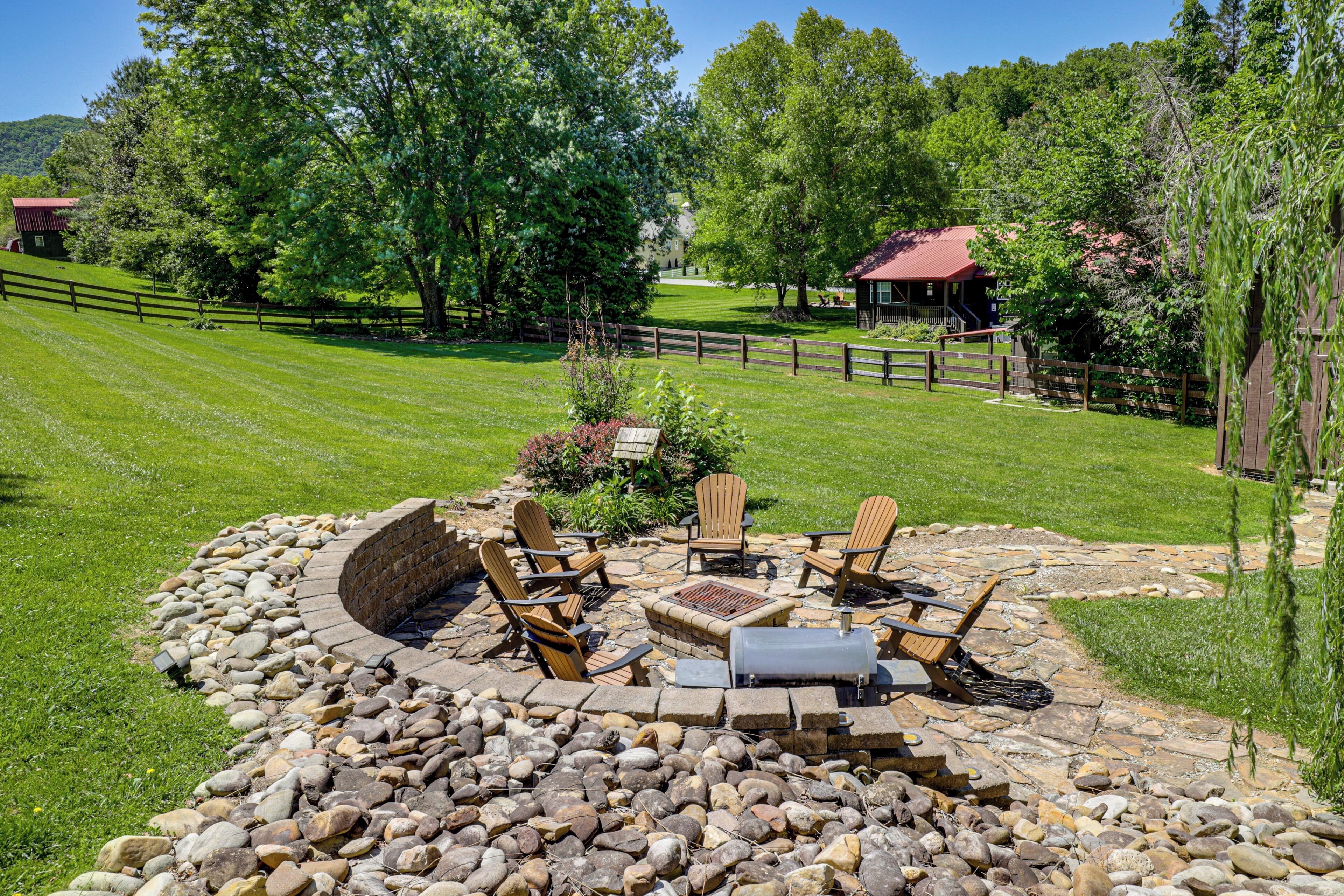 A stone fire pit with outdoor furniture, surrounded by a stone wall, in a grassy area with trees and a wooden fence in the background.