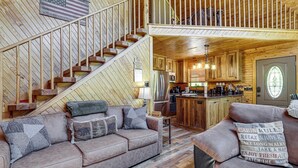 A cabin interior with a wooden staircase, a kitchen area, a living room with a sofa, and a second-floor balcony. - Views, Fire Pit + Game Room: Smoky Mountain Cabin (Sevierville)