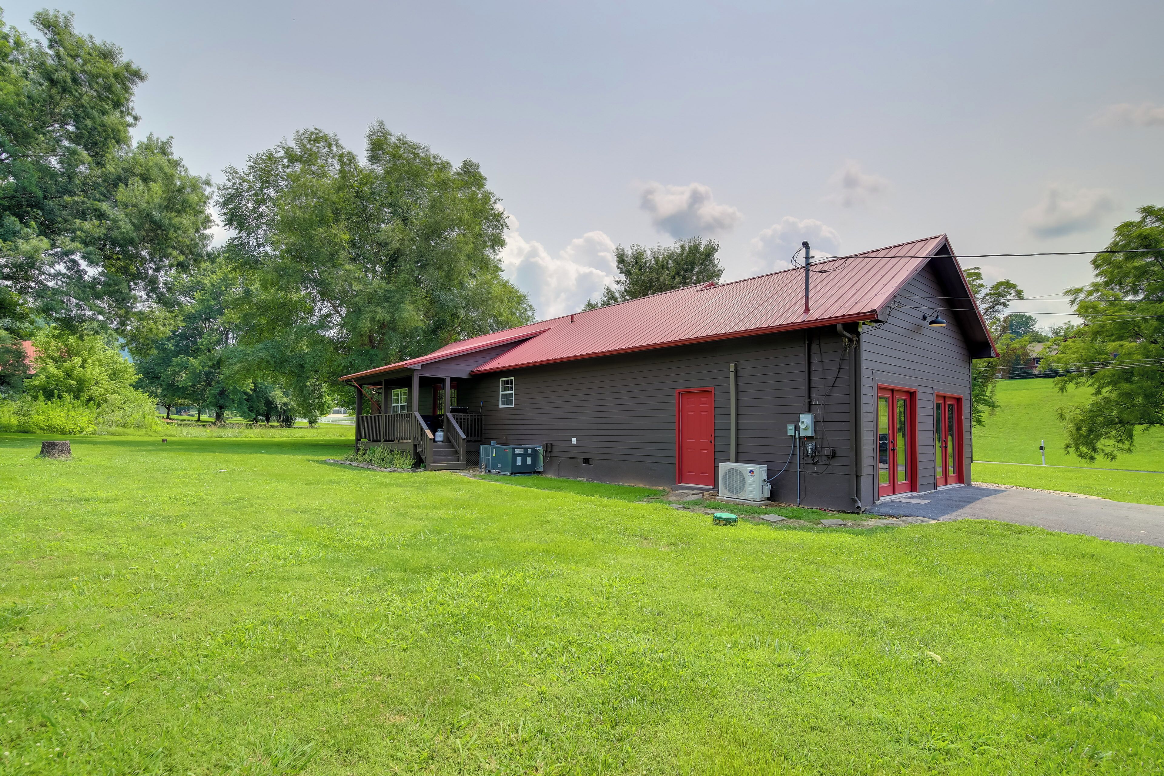 A single-story building with a red roof and a red door, surrounded by a grassy field and trees.