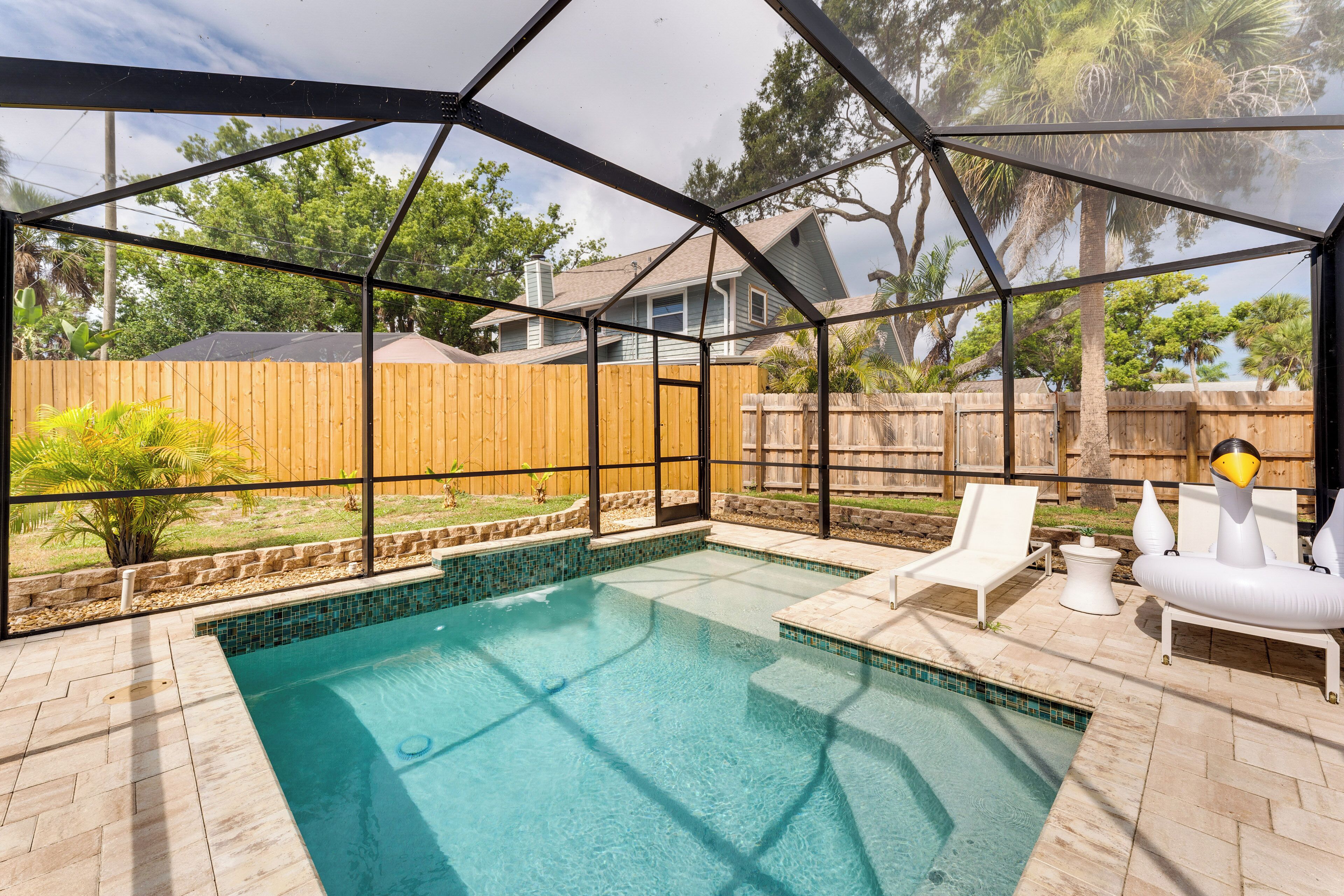 A covered outdoor swimming pool with a wooden fence, lounge chairs, and a view of a house in the background.