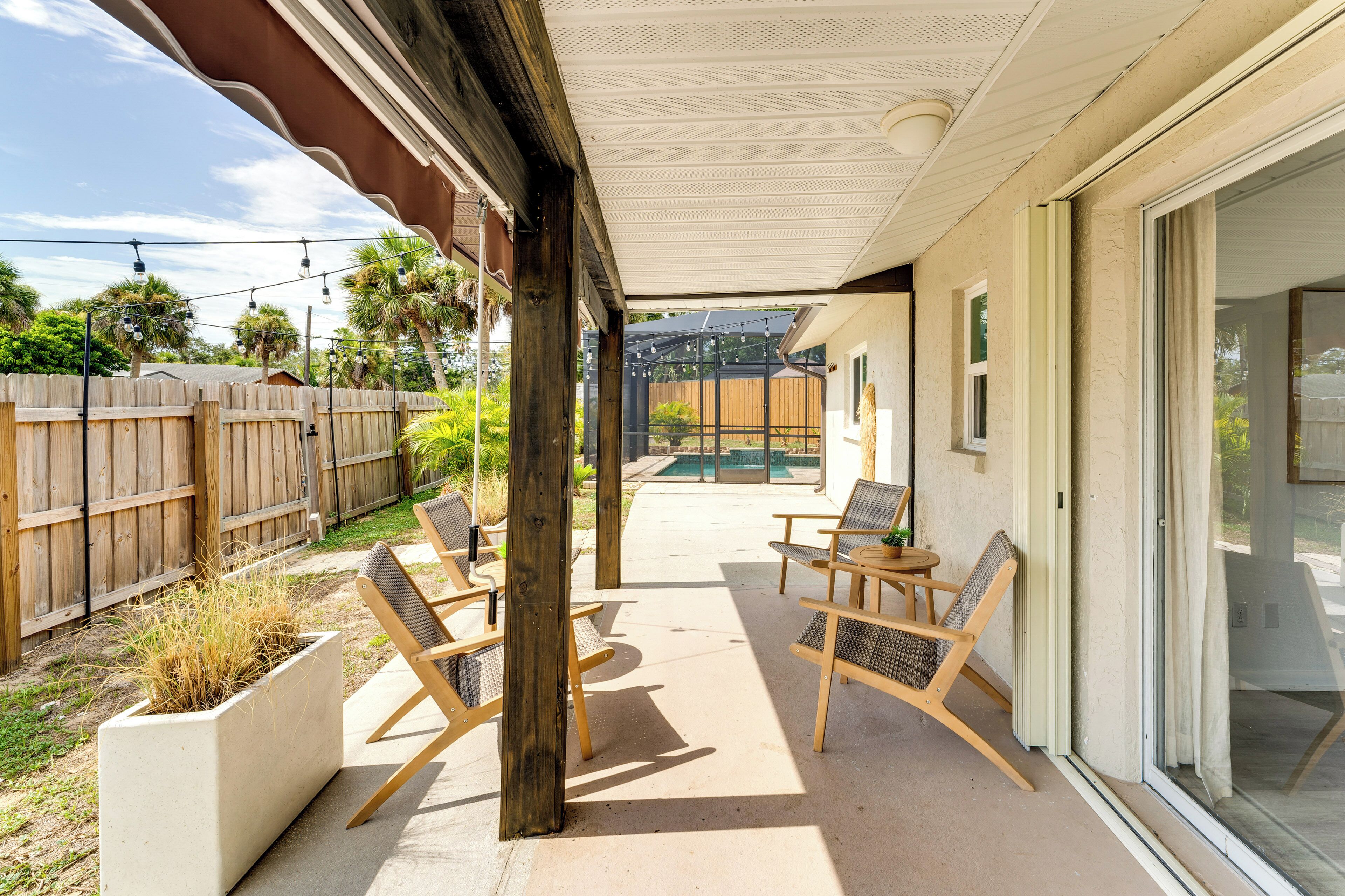 A covered patio area with wooden chairs and a concrete floor, overlooking a backyard with a wooden fence and a pool.