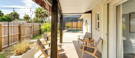 A covered patio area with wooden chairs and a concrete floor, overlooking a backyard with a wooden fence and a pool.
