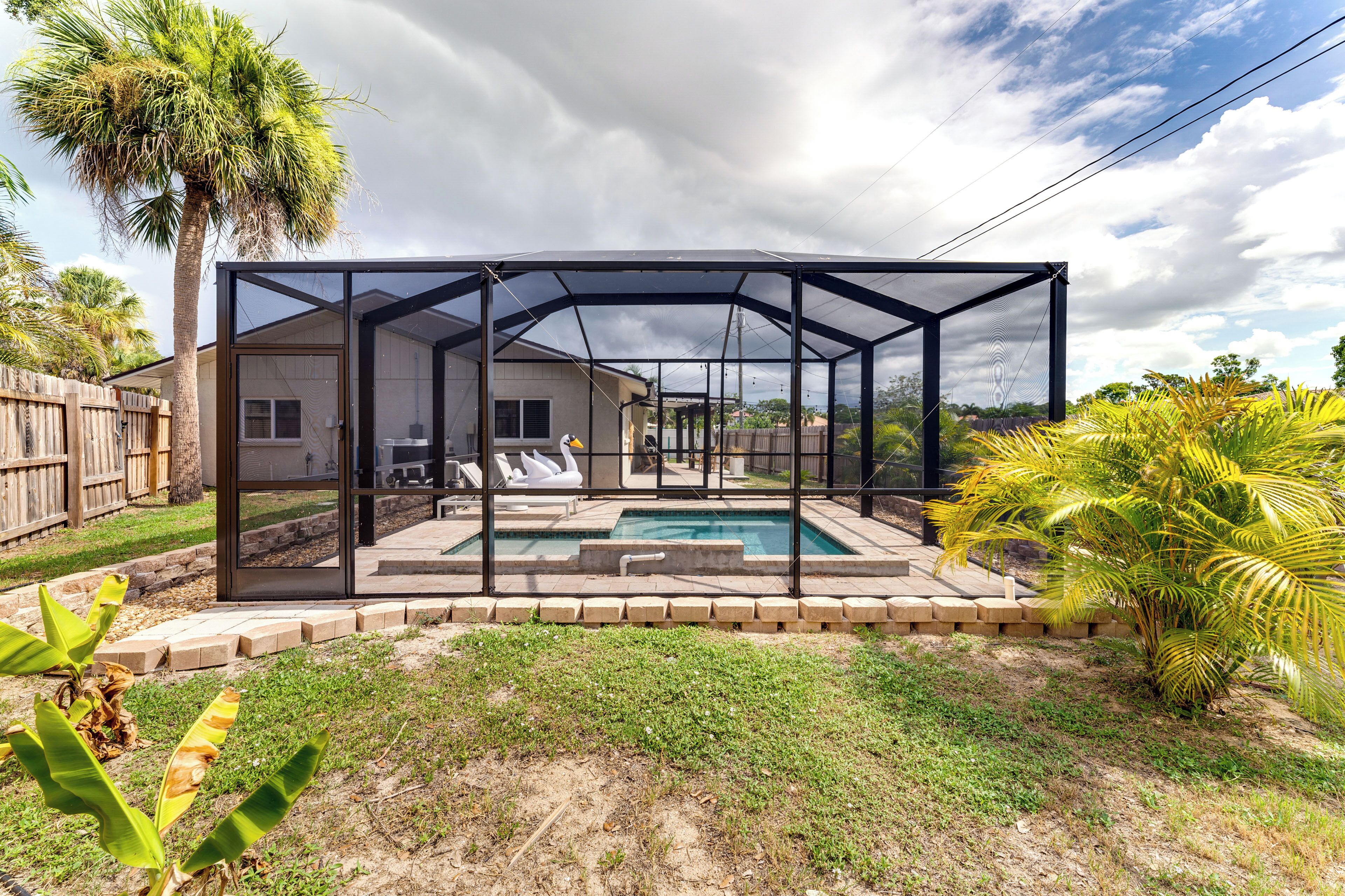 A covered outdoor swimming pool with a glass enclosure, surrounded by a brick border, palm trees, and a wooden fence.