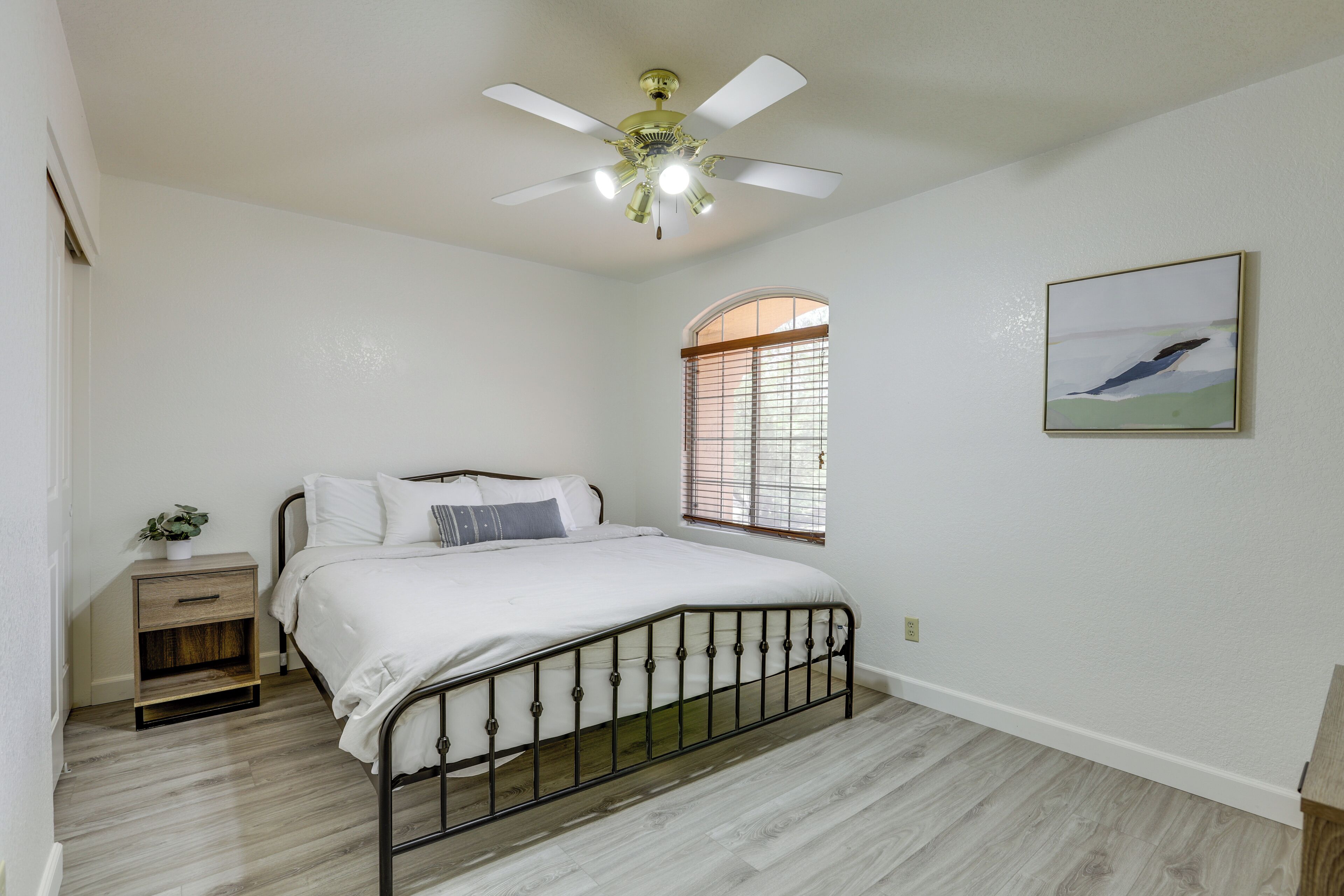 A bedroom with a bed, a nightstand, a ceiling fan, a window with blinds, and a painting on the wall.