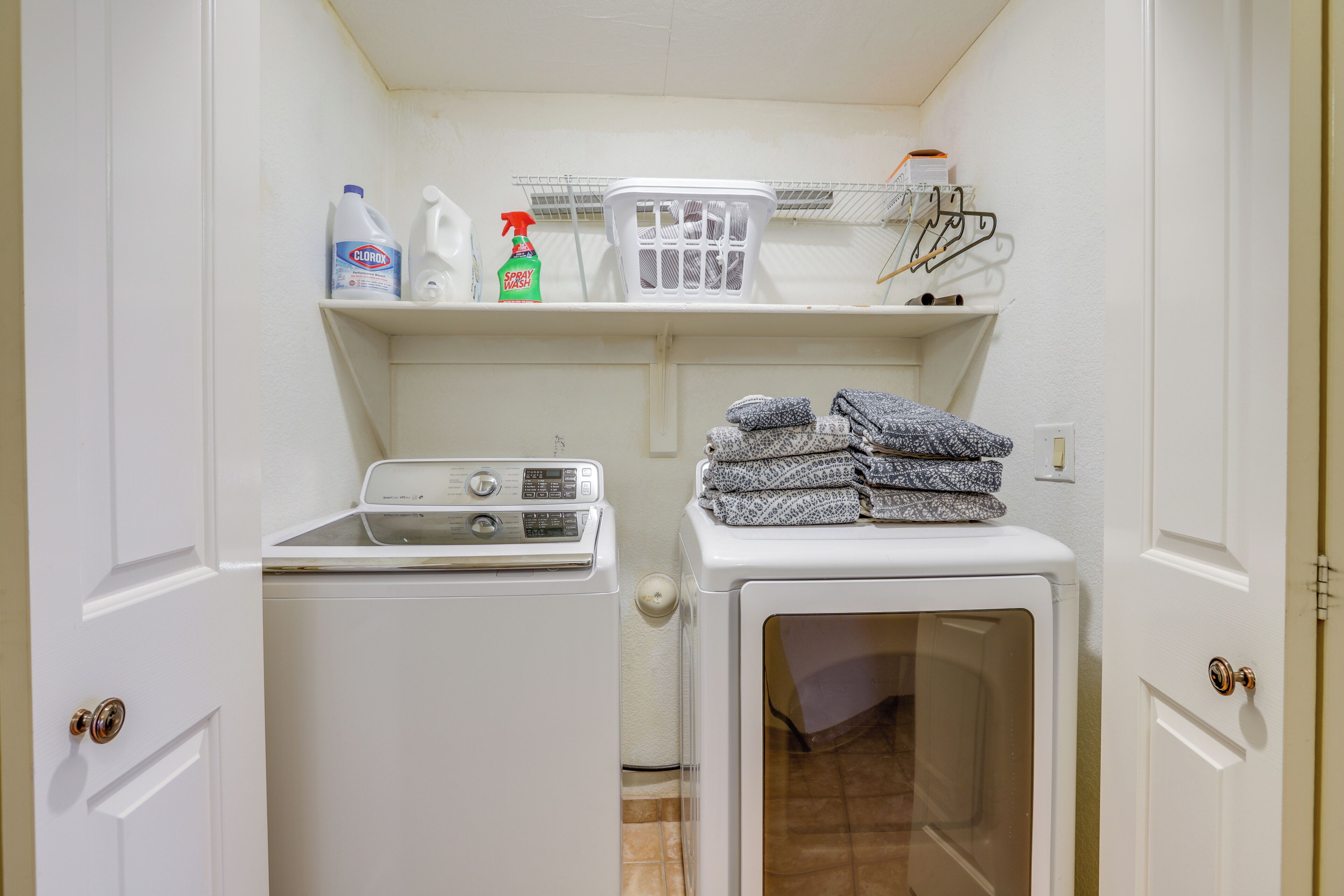 A laundry room with a front-loading washer and dryer, a shelf with cleaning supplies, and a basket.