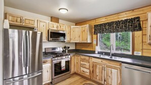 A modern kitchen with wooden cabinets, stainless steel appliances, and a large window with a view of trees. - Welcoming Wasilla Cabin w/ Patio! (Wasilla)
