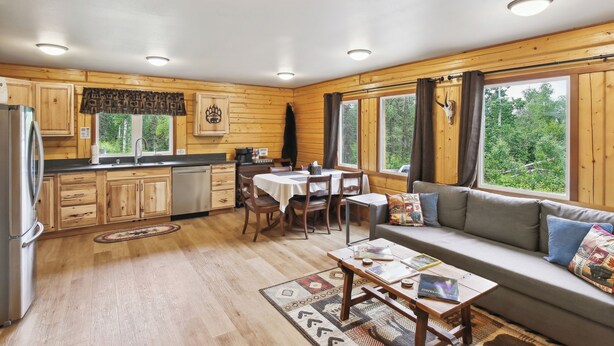 A cabin interior with wooden walls, a dining area, a sofa, and a kitchen with a refrigerator and cabinets. - Welcoming Wasilla Cabin w/ Patio! (Wasilla)