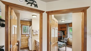 A cabin interior with wooden walls, a kitchen area with a refrigerator, a sink, and a window. There is a chair and a bed visible in the room. - Welcoming Wasilla Cabin w/ Patio! (Wasilla)