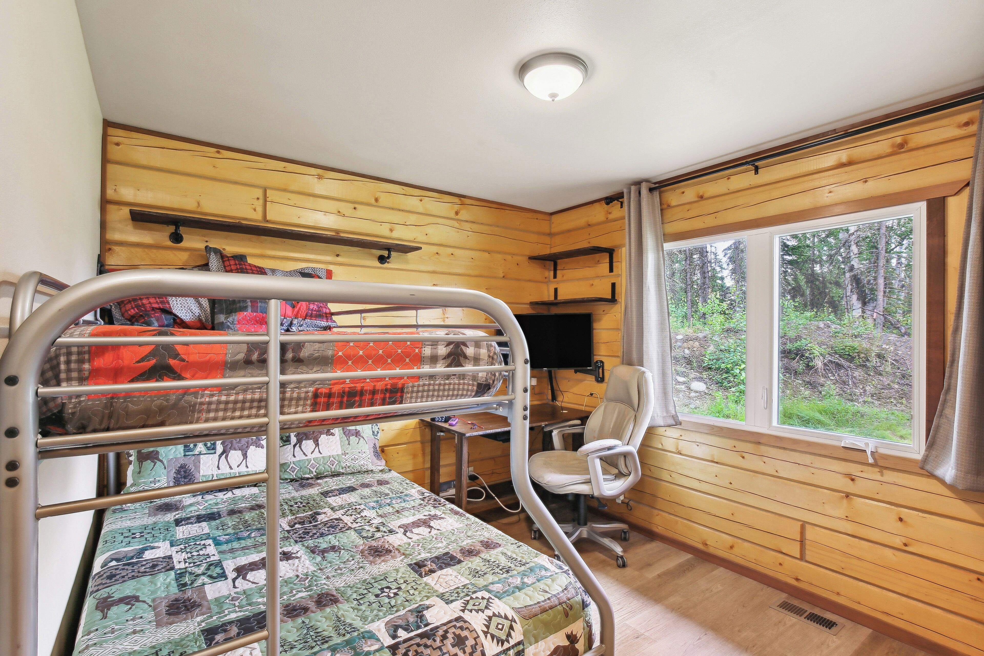Ein Etagenbett mit Steppdecke, ein Schreibtisch mit Stuhl, ein Fernseher und ein Fenster mit Blick auf einen Wald.