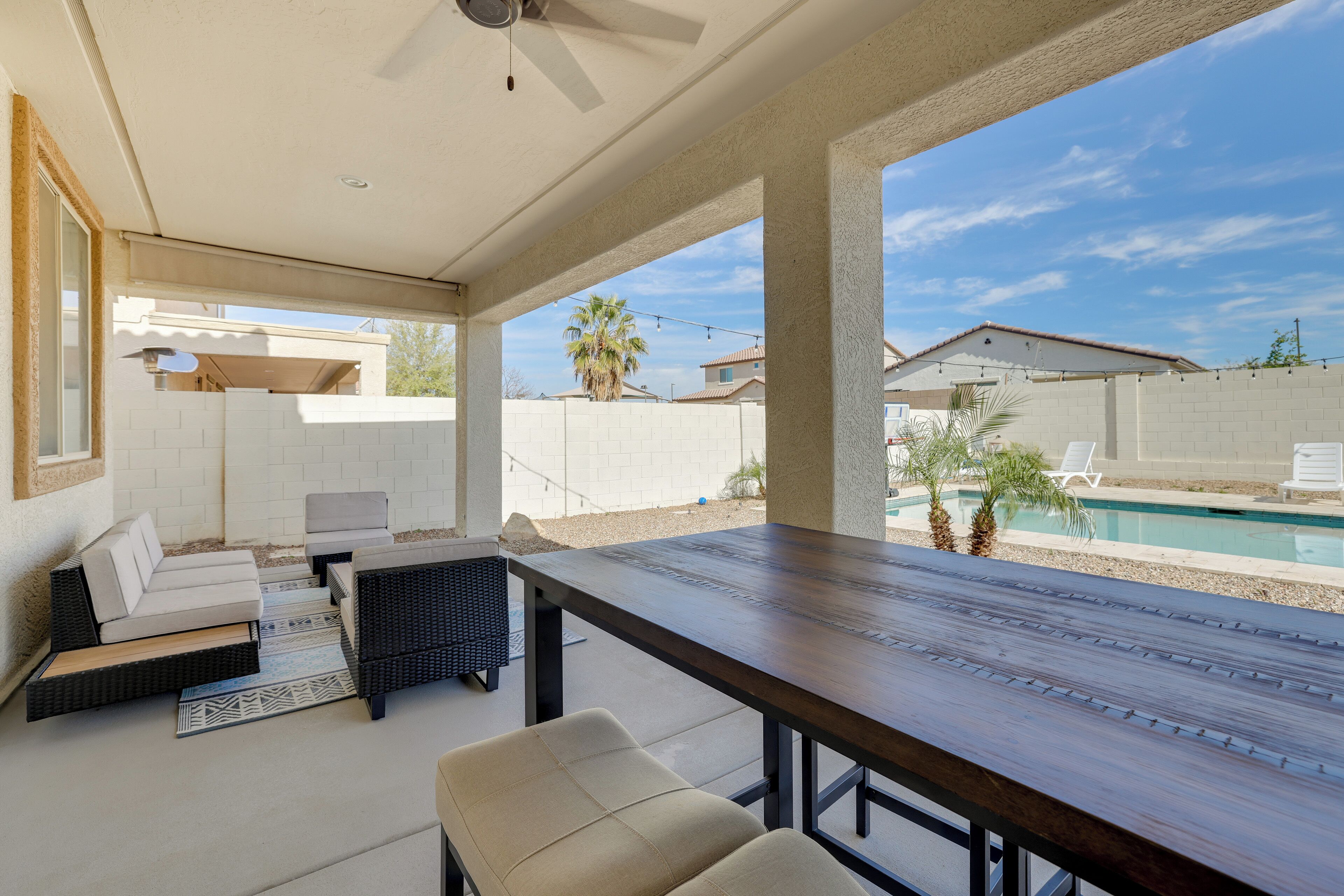 Une terrasse extérieure avec une table et des chaises, un ventilateur de plafond, et une vue sur une piscine et les maisons voisines.