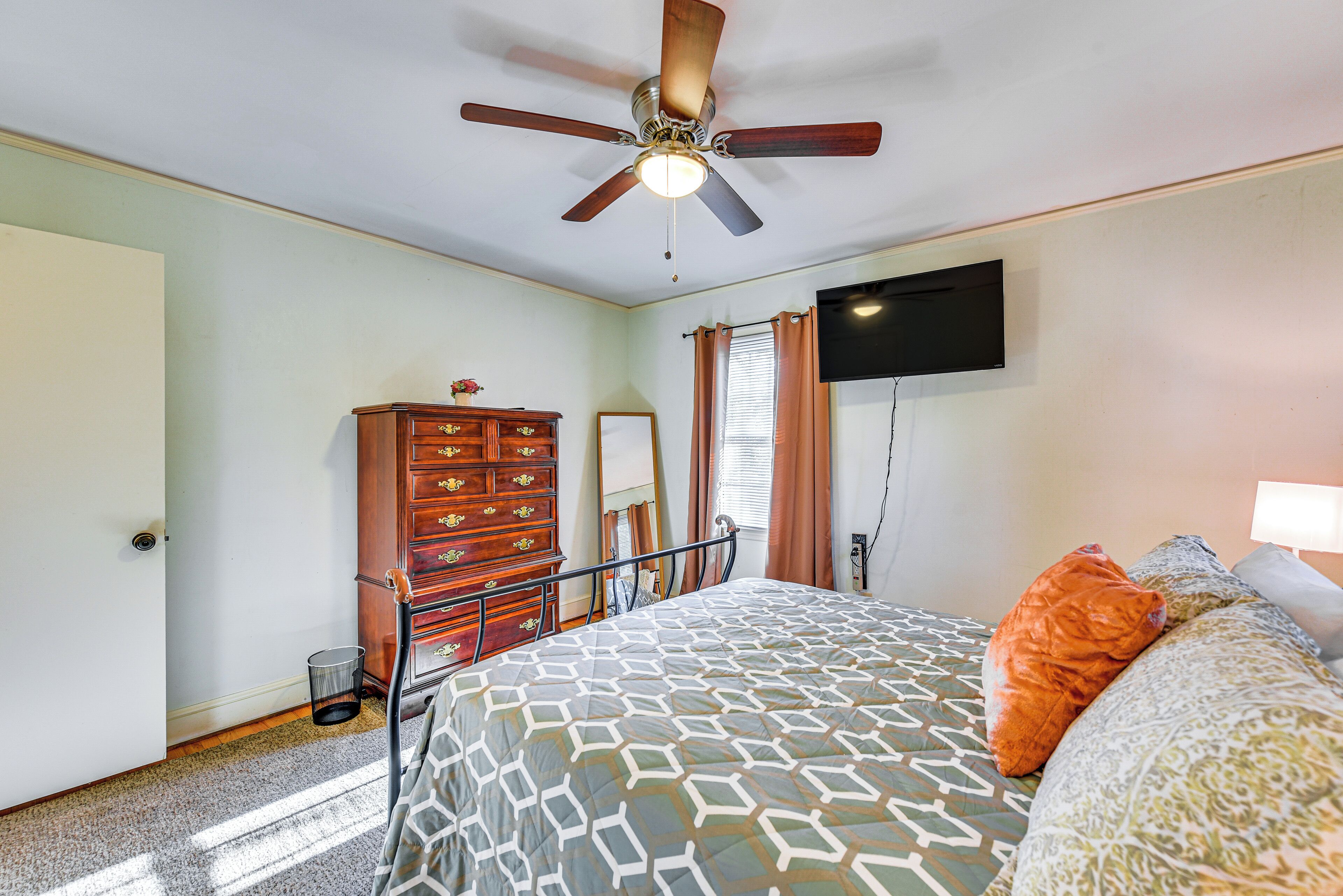 Un dormitorio con cama, ventilador de techo, cómoda, espejo y televisor montado en la pared.