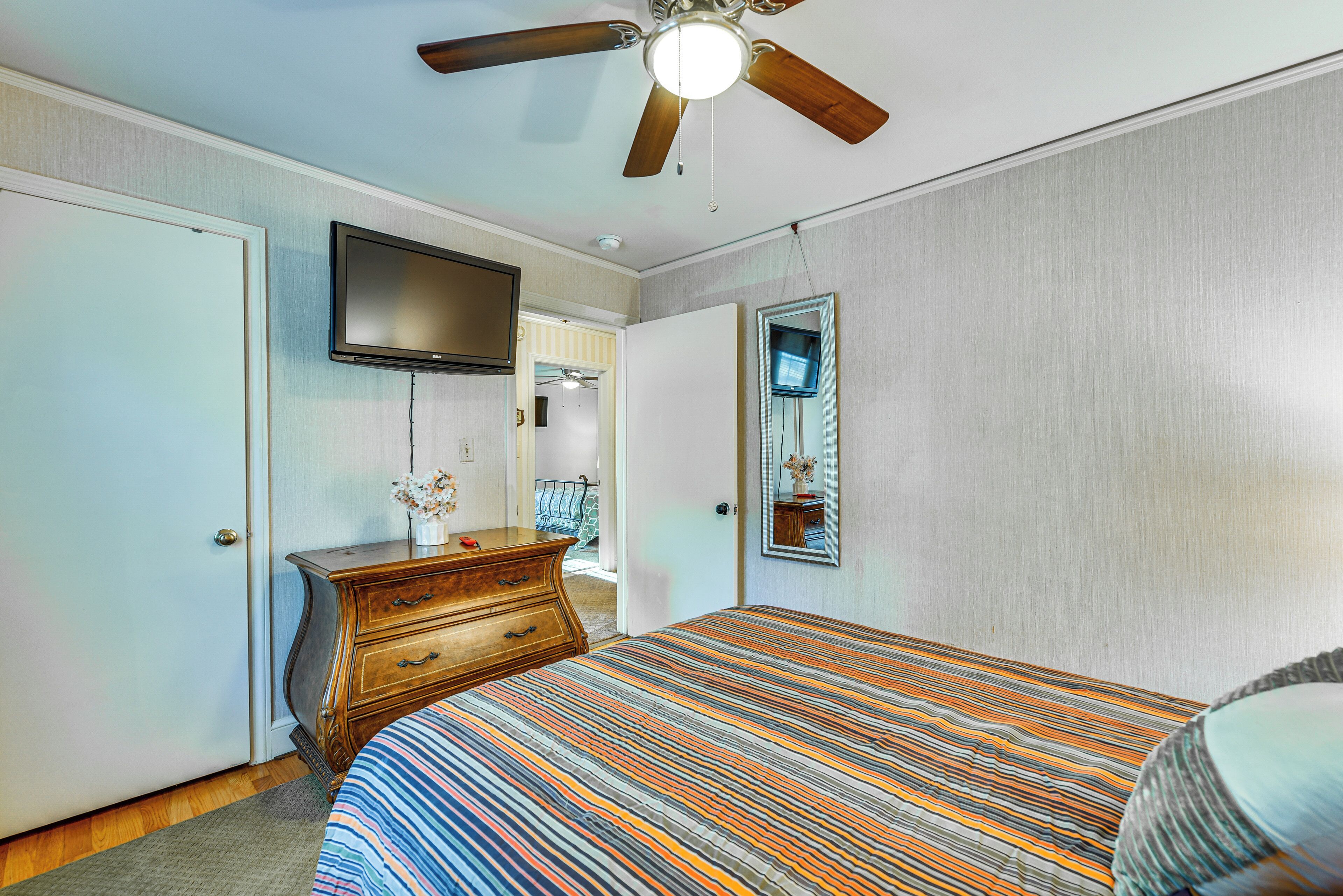 A bedroom with a bed, dresser, TV, ceiling fan, and a mirror.
