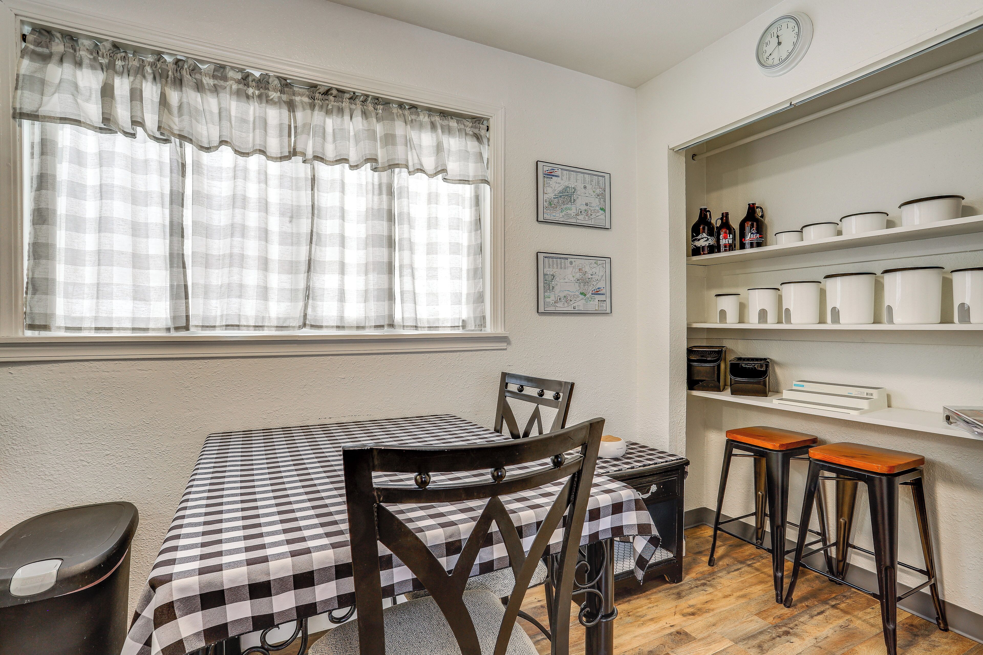 A dining area with a checkered table, two barstools, a window with curtains, and shelves with kitchenware.