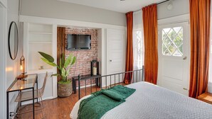 A bedroom with a bed, a TV mounted on a brick wall, a desk with a chair, and a window with curtains. - 1 Mi to Dtwn! Bright Long Beach Apartment (Long Beach)
