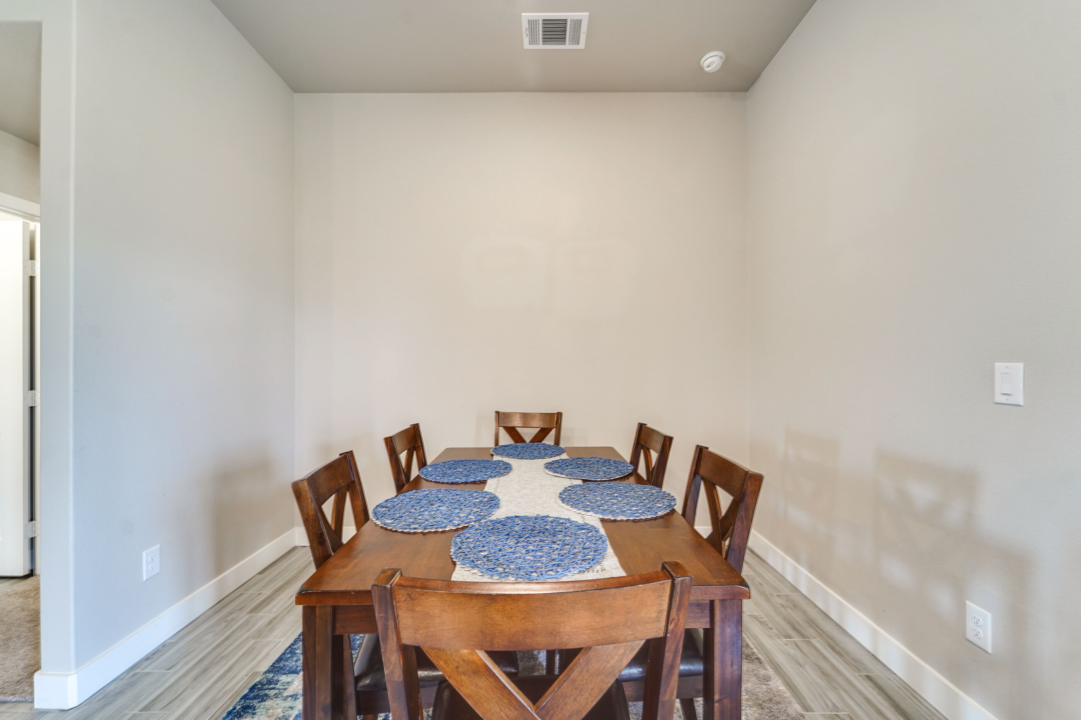Un comedor con una mesa de madera y seis sillas, una alfombra en el centro y una pared lisa.