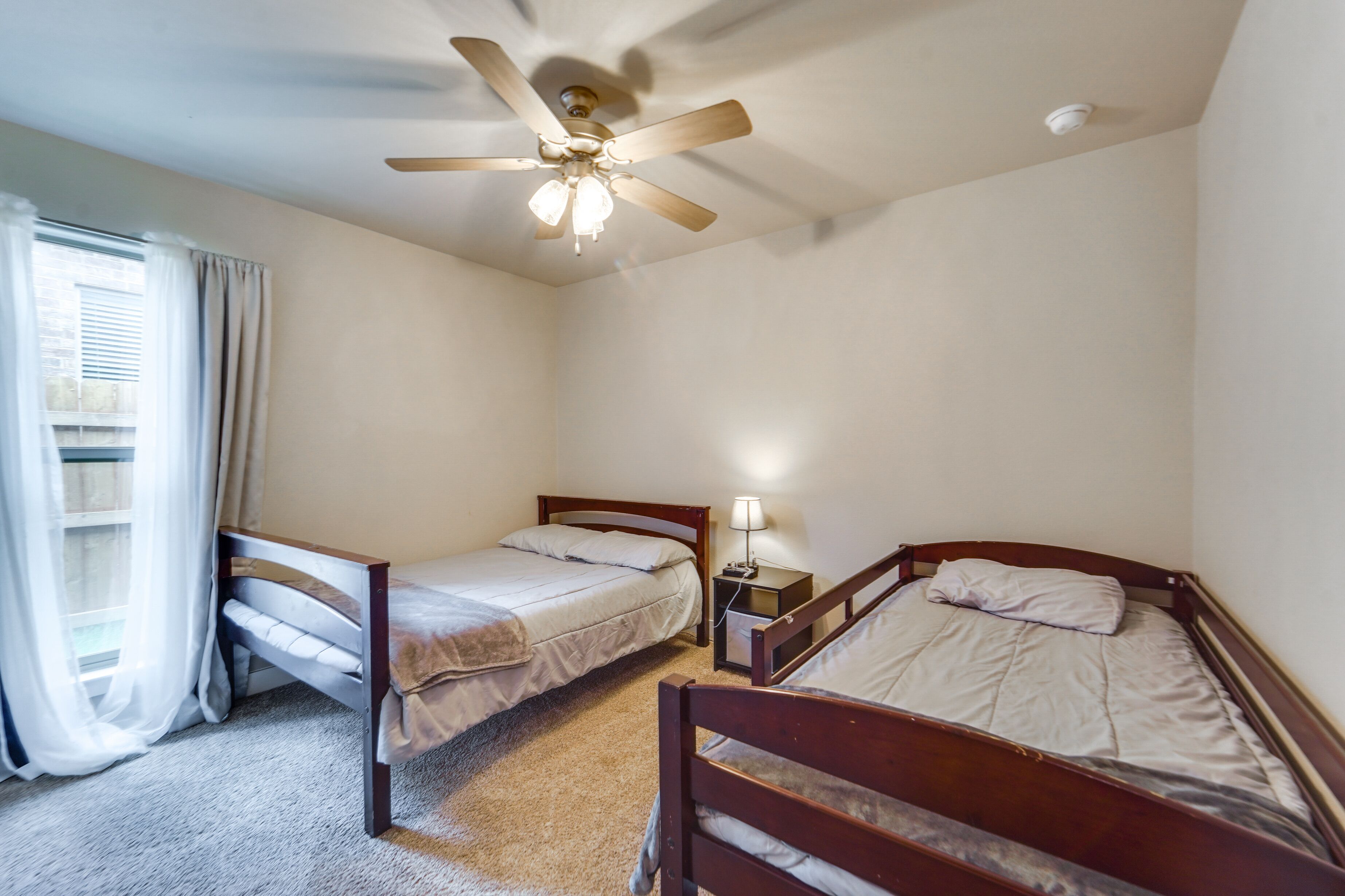 A bedroom with two single beds, a ceiling fan, a nightstand with a lamp, and a window with curtains.