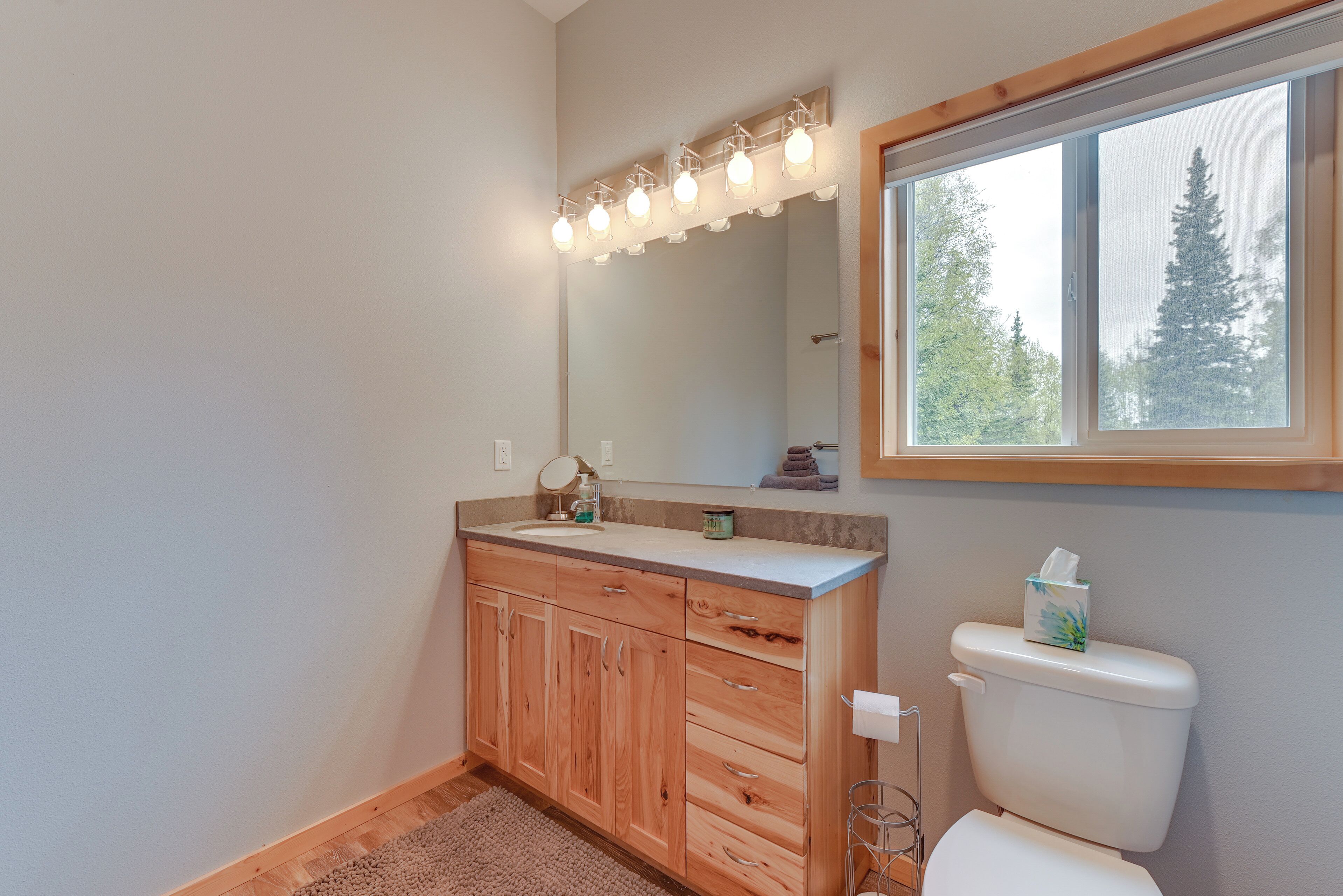 A bathroom with wooden cabinets, a sink, a mirror with lights, and a window with a view of trees.