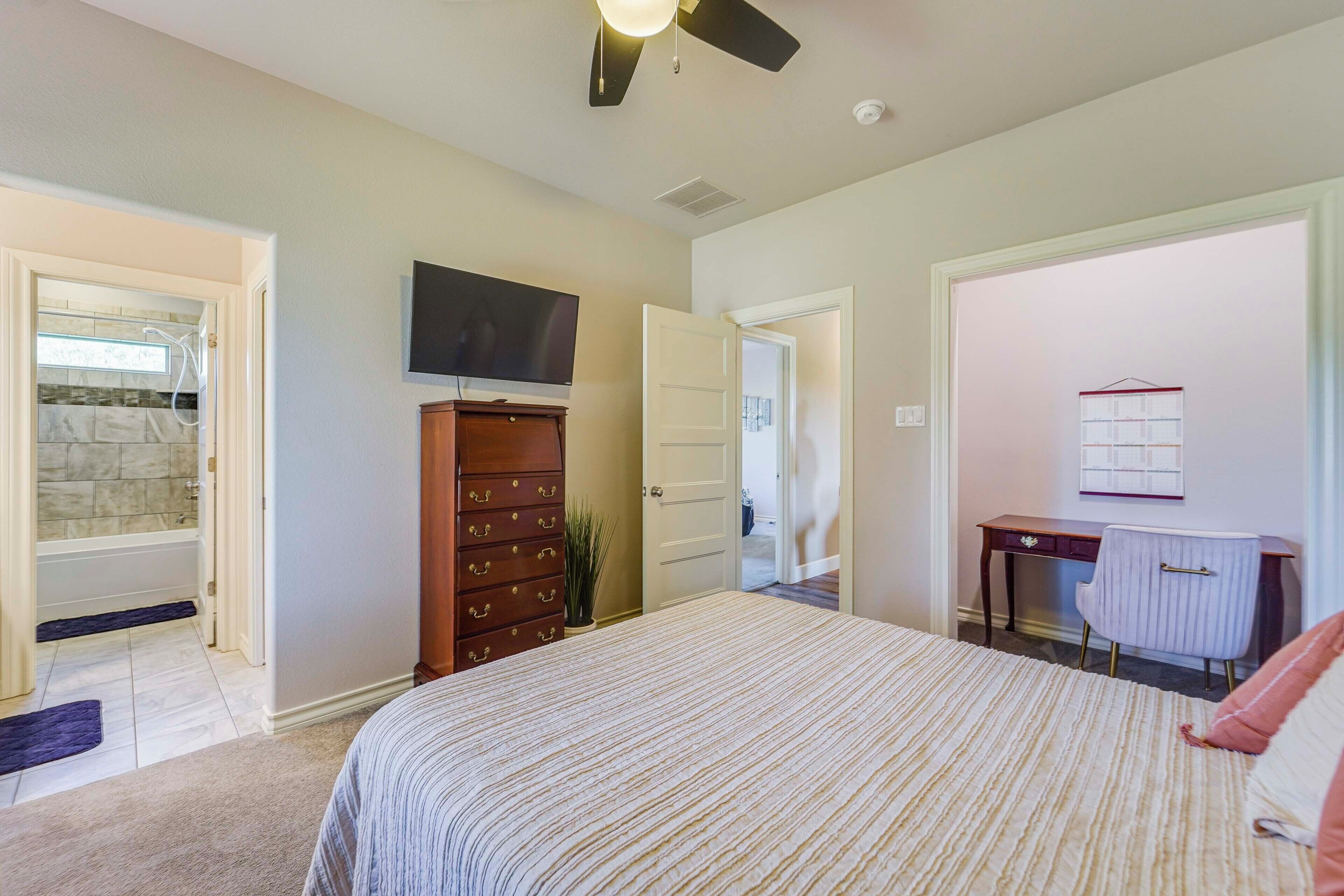 A bedroom with a bed, dresser, TV, desk, chair, and a door leading to a bathroom.