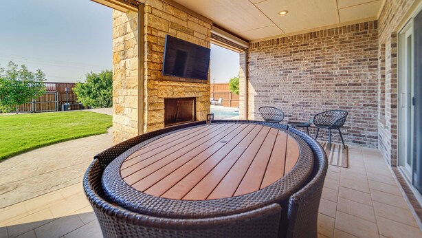An outdoor patio with a round wooden table and wicker chairs, a stone fireplace, and a flat-screen TV mounted on the wall. - Spacious Lubbock Home w/ Private Pool & Yard! (Lubbock)