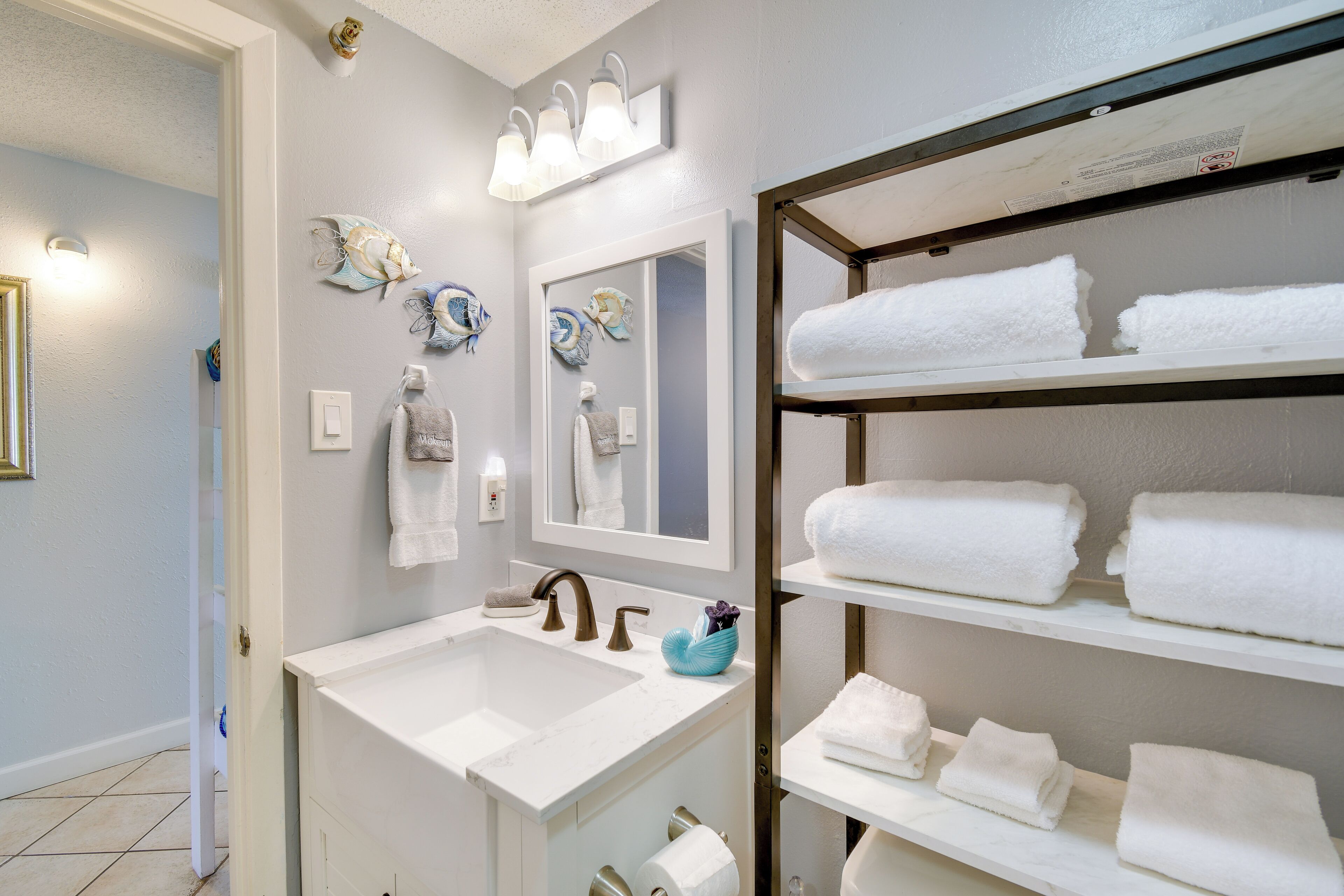A bathroom with a white sink, a mirror, decorative wall art, and shelves with folded towels.