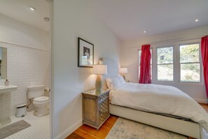 Une chambre à coucher comprenant un lit, des lampes de chevet, une table de chevet, une fenêtre avec des rideaux, et une salle de bain avec des toilettes et un lavabo.
