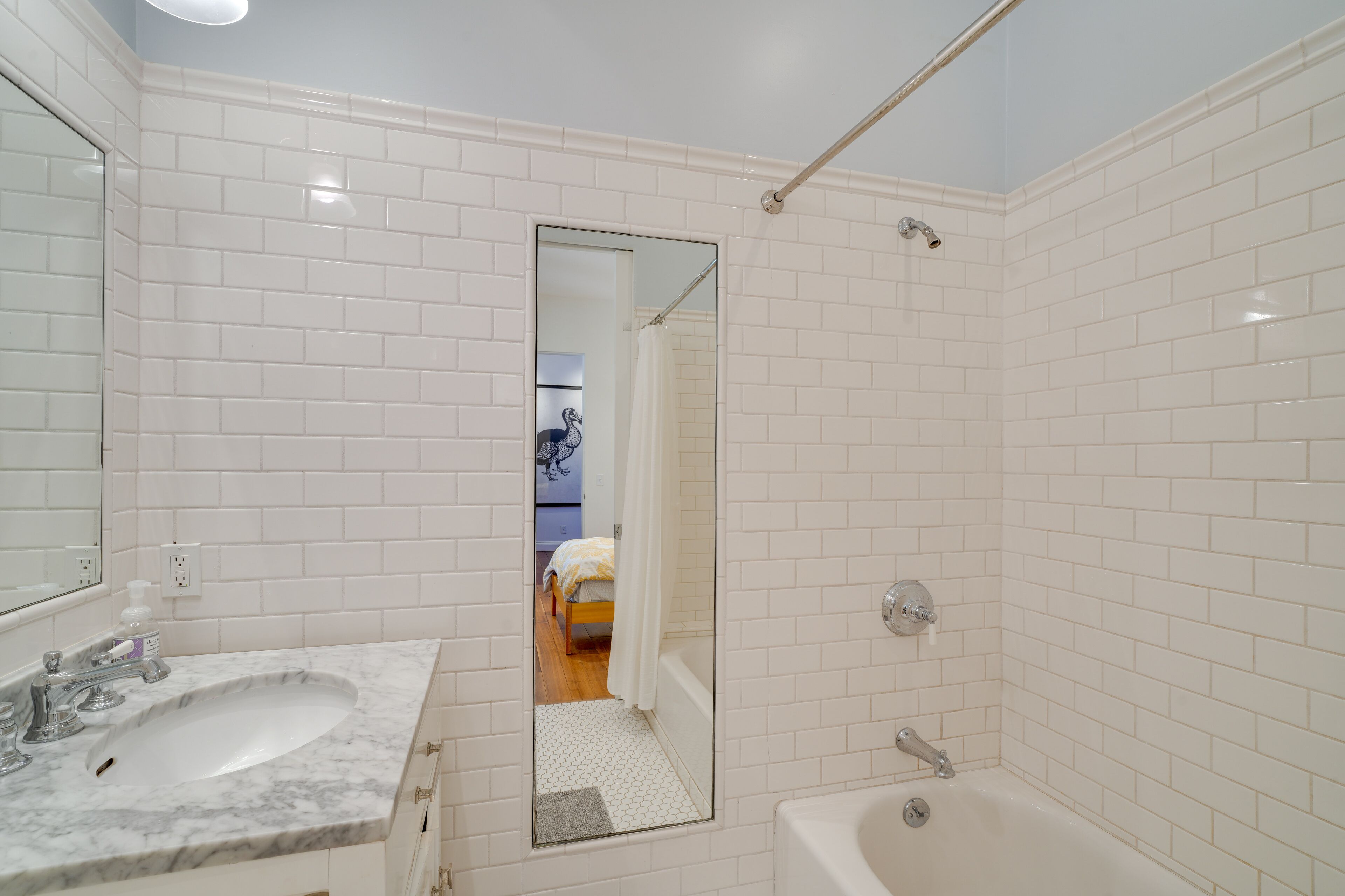 A bathroom with white tiled walls, a marble sink, a bathtub, and a mirror reflecting a bedroom with a bed and a shower curtain.