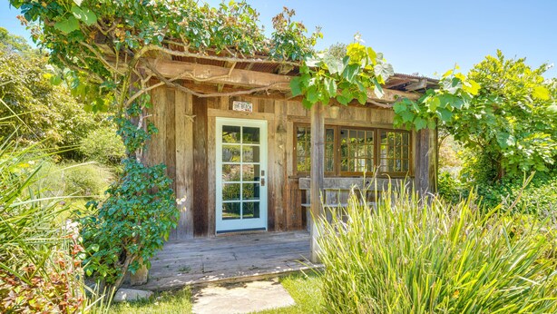 A rustic wooden cabin with a white door, surrounded by lush greenery and a wooden deck. - Farmhouse-chic Carpinteria Cottage w/ Pool Access (Carpinteria)