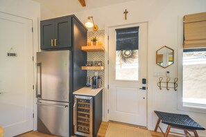 A compact kitchen with a built-in refrigerator, a coffee machine, and a wine cooler. There is a small shelf with potted plants and a window with a view of palm trees.