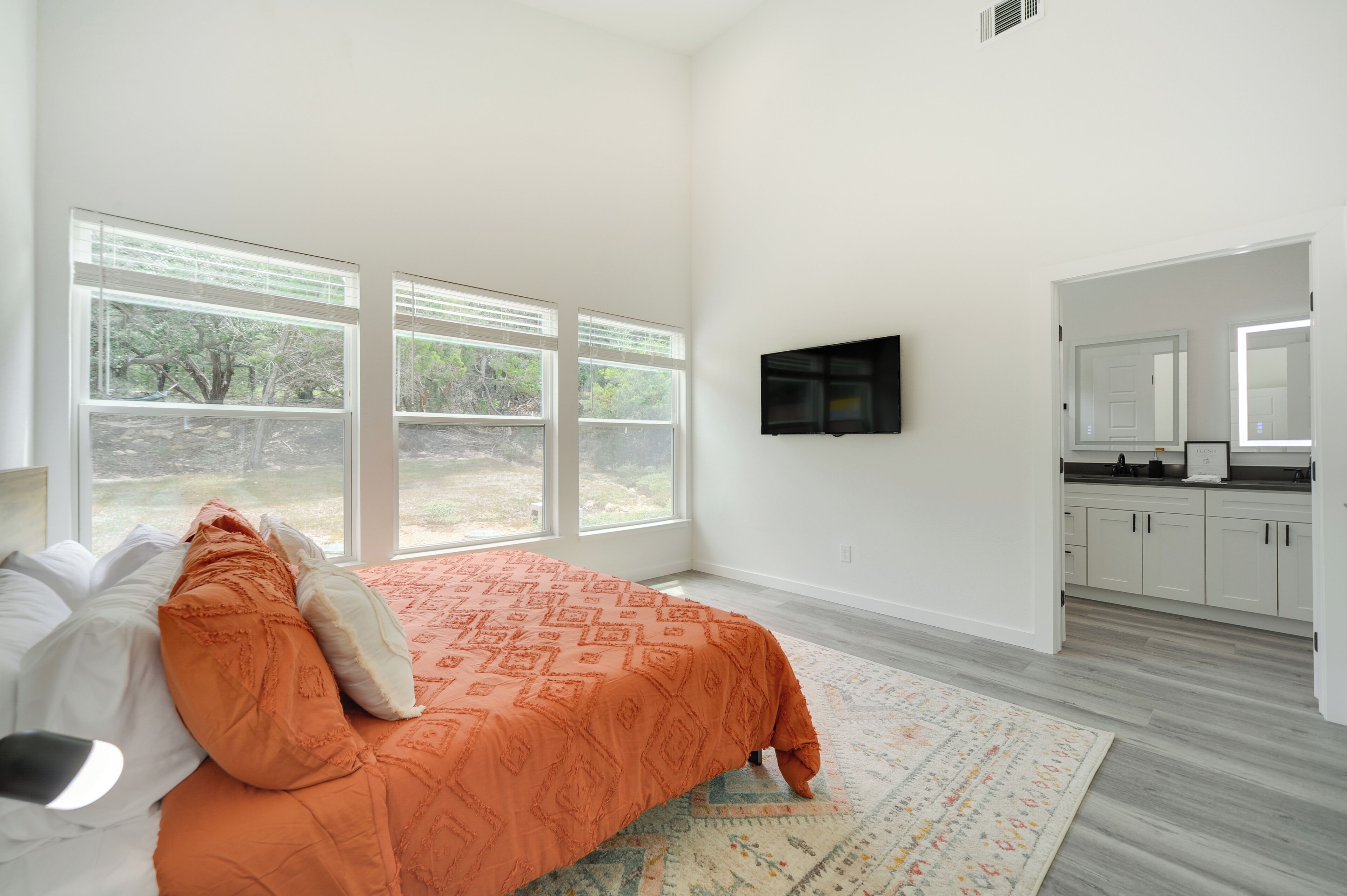 A bedroom with a large bed, a TV mounted on the wall, and a view of the outdoors through large windows.